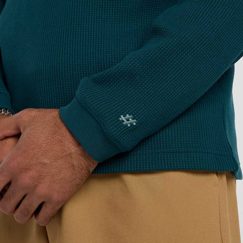 A close-up shows a person wearing the New Balance Numeric Waffle Knit Tee MT51958AAV in textured teal, featuring a small white stitched logo on the cuff, paired with tan pants. Their hands are clasped in front.