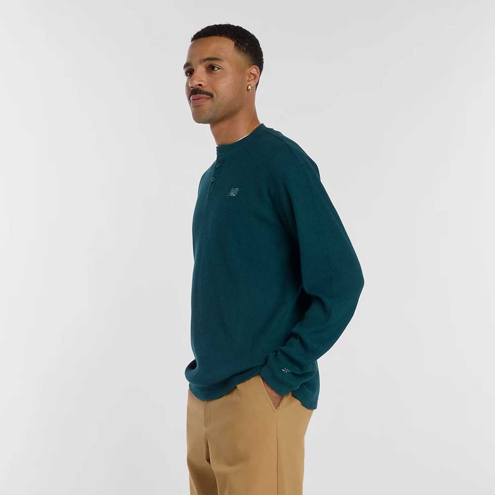 A man with short hair and a mustache stands sideways, hands in his pockets, wearing the New Balance Numeric Waffle Knit Tee MT51958AAV in teal and tan pants against a plain white background.