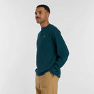 A man with short hair and a mustache stands sideways, hands in his pockets, wearing the New Balance Numeric Waffle Knit Tee MT51958AAV in teal and tan pants against a plain white background.