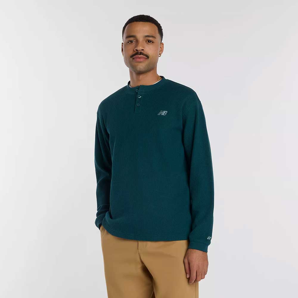 A man wearing a teal New Balance Numeric Waffle Knit Tee MT51958AAV and tan pants stands against a plain white background, looking at the camera with a neutral expression.