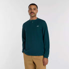 A man wearing a teal New Balance Numeric Waffle Knit Tee MT51958AAV and tan pants stands against a plain white background, looking at the camera with a neutral expression.