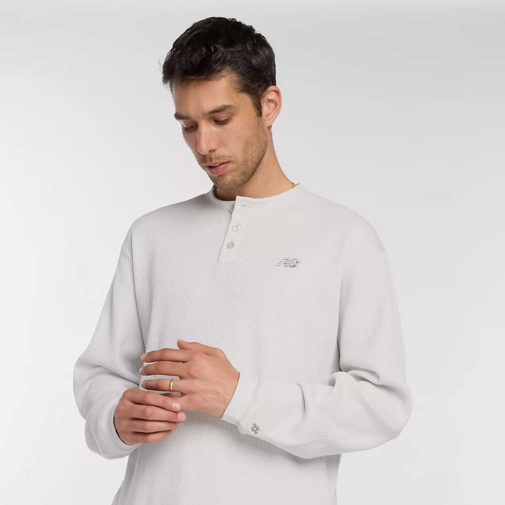 A man with short brown hair wearing the New Balance Numeric Waffle Knit Tee MT51958GYM in light gray looks down while touching his hands against a plain, light-colored background.