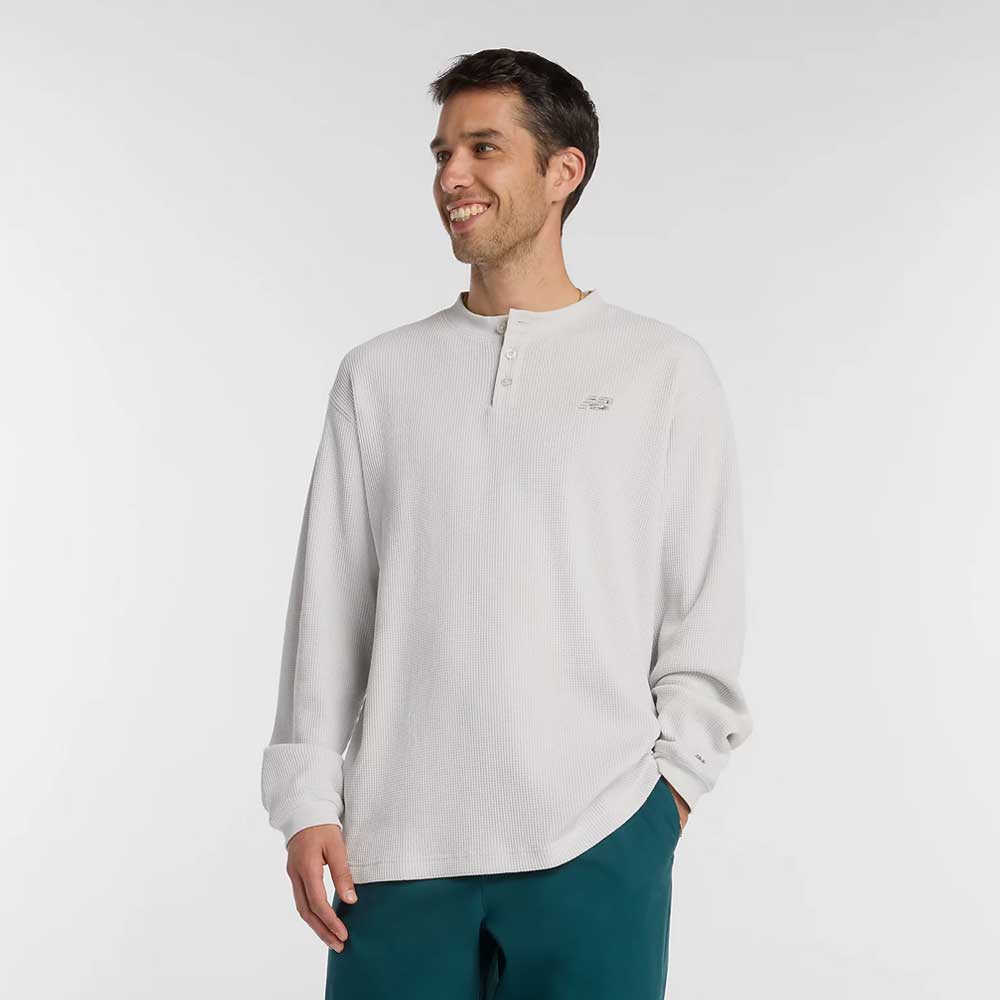 A man wearing the New Balance Numeric Waffle Knit Tee MT51958GYM in light gray and teal pants stands smiling against a plain white background.