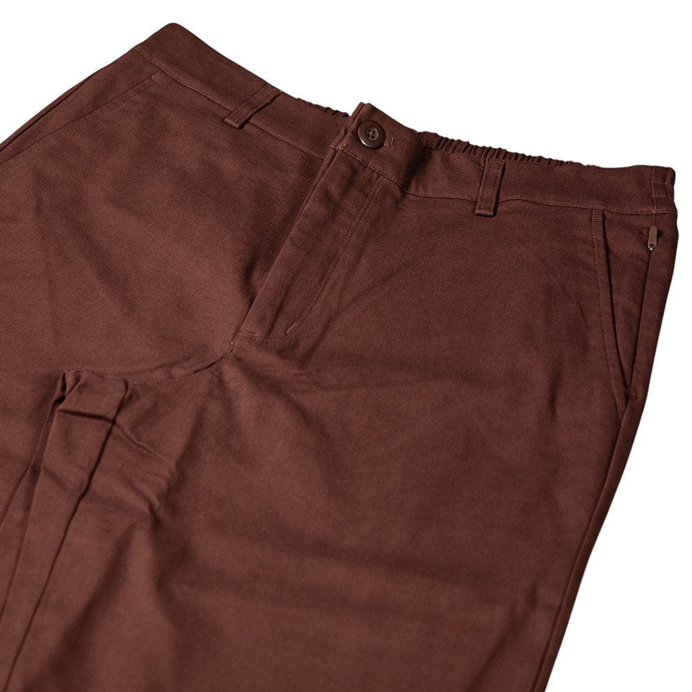 The New Balance Numeric Pant Pumperknickel MP51953ABJ is a brown cotton canvas pant with belt loops, button closure, side pockets, and an elastic waistband on one side, shown laid flat on a white surface.