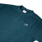 The New Balance Numeric Waffle Knit Tee MT51958AAV is a dark teal short-sleeve henley shirt with a textured waffle knit, three collar buttons, and a small white embroidered logo on the left chest, shown on a white background.