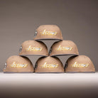 Six Chalino Skull Camel Storm Houston Astros New Era Hat 5950 Fitted caps are stacked in a pyramid on a light surface, each showing “Astros” in gold. The neutral gray gradient background highlights their sleek design.