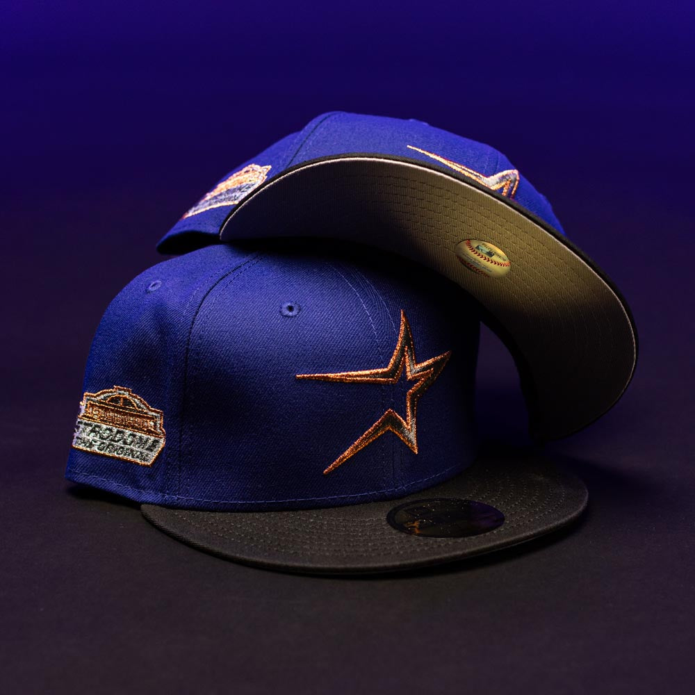 Two blue and black H Town North Houston Astros New Era Hat 5950 Fitted caps in OG Dark Royal, each adorned with gold star logos and World Series Championship patches for the Houston Astros, displayed on a dark surface against a purple background.