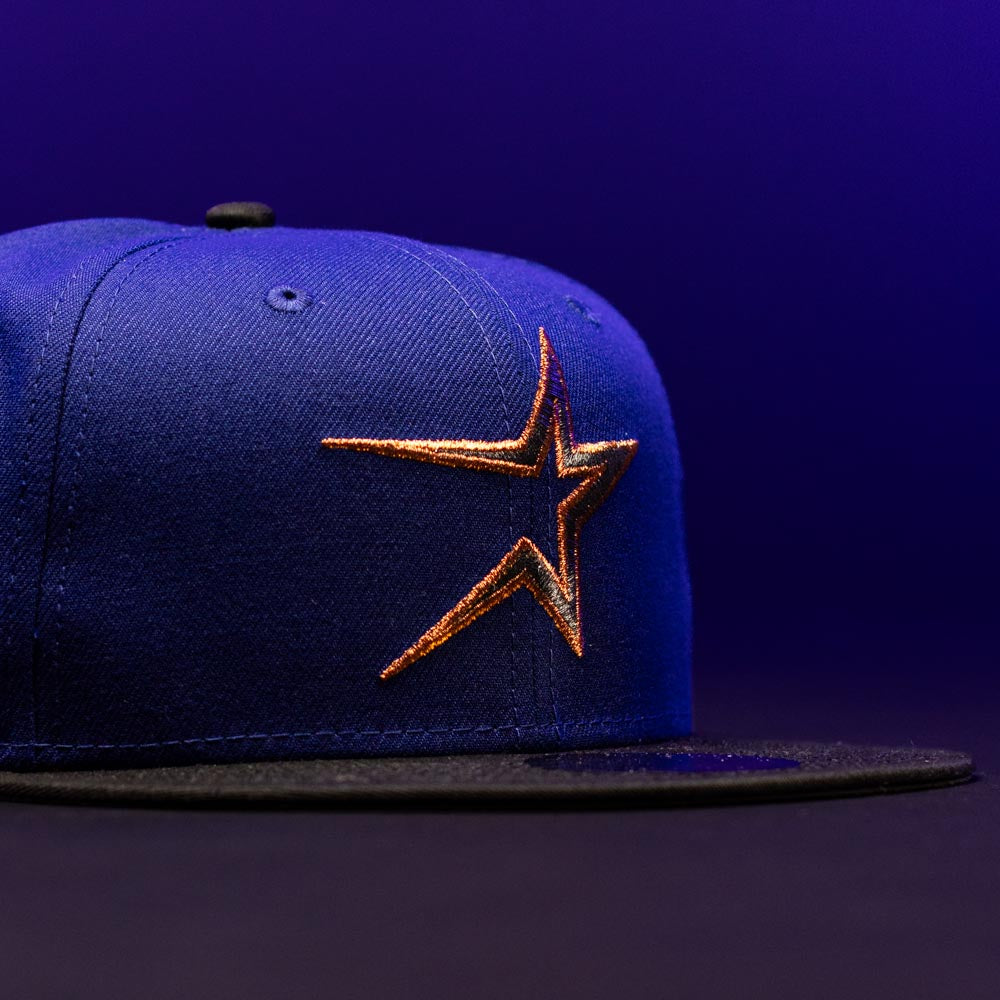 The H Town North Houston Astros New Era Hat 5950 Fitted features a blue crown with a black brim and showcases an orange and gold Astros star logo on the front, set against a dark purple background. This OG Dark Royal design by New Era makes a striking addition to your collection.