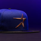 The H Town North Houston Astros New Era Hat 5950 Fitted features a blue crown with a black brim and showcases an orange and gold Astros star logo on the front, set against a dark purple background. This OG Dark Royal design by New Era makes a striking addition to your collection.