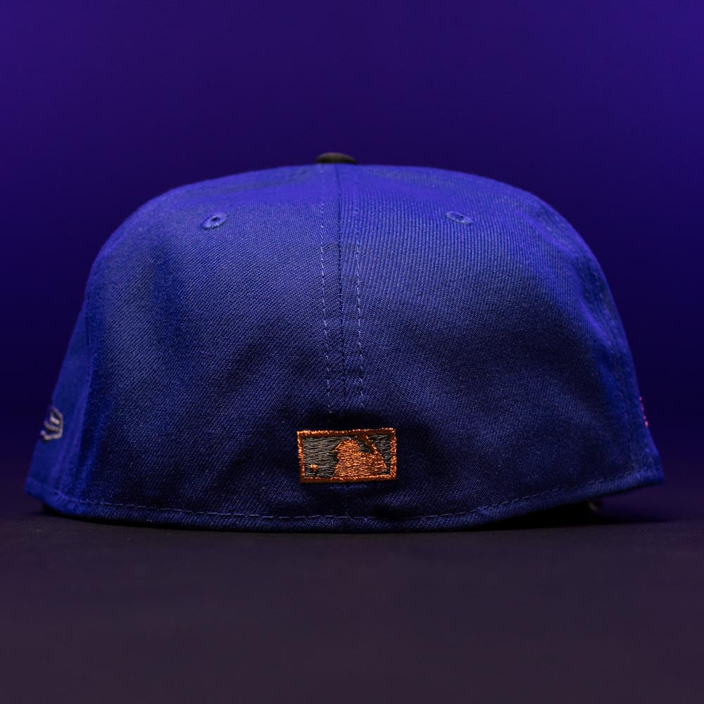 A blue H Town North Houston Astros New Era Hat 5950 Fitted is displayed from the back against a purple background, featuring a small brown and black stitched logo patch near the bottom center. The OG Dark Royal color adds a bold touch, making this cap stand out in any collection.