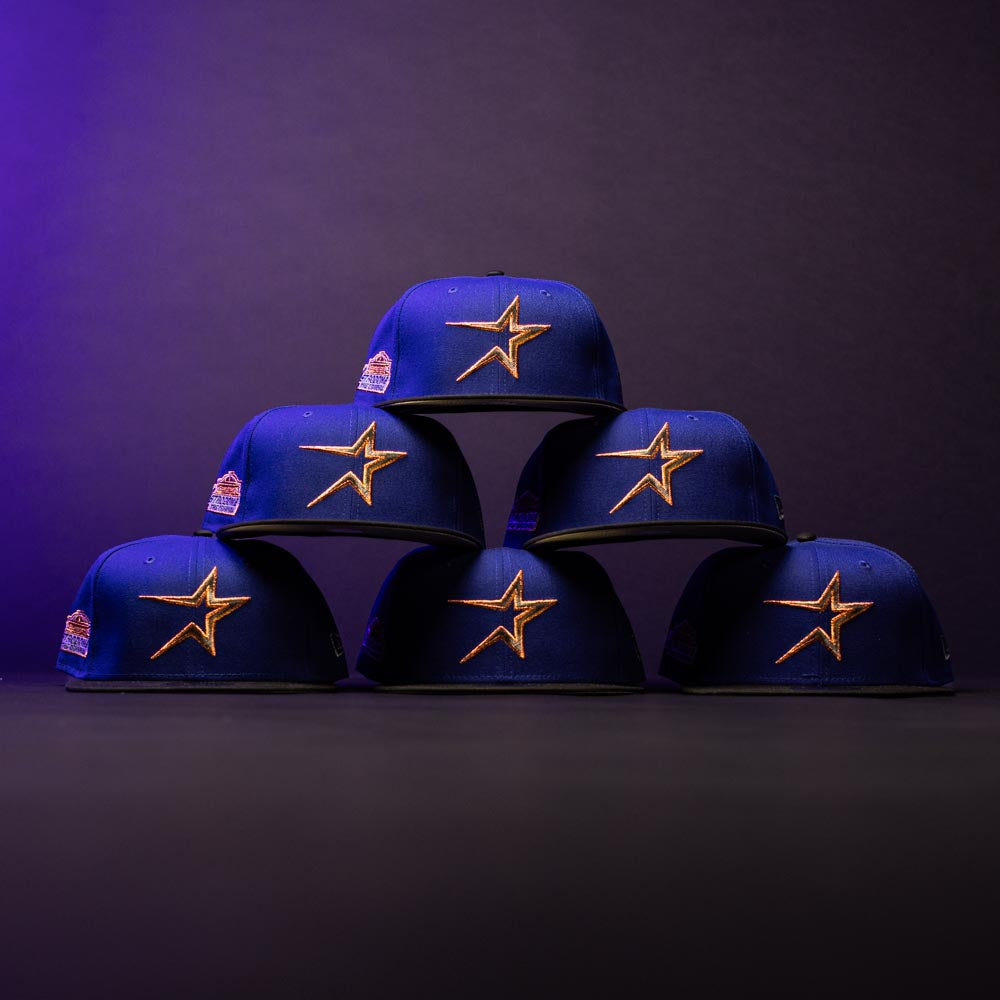 Six H Town North Houston Astros New Era Hat 5950 Fitted caps, featuring an OG Dark Royal blue colorway and gold star logos, are stacked in a pyramid on a dark surface against a gradient purple-black background.