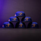 Six H Town North Houston Astros New Era Hat 5950 Fitted caps, featuring an OG Dark Royal blue colorway and gold star logos, are stacked in a pyramid on a dark surface against a gradient purple-black background.