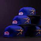 Four H Town North Houston Astros New Era Hat 5950 Fitted caps, featuring OG Dark Royal blue fabric, gold star embroidery, and a colorful Houston Astros patch, are stacked in a pyramid on a dark surface with dramatic lighting.