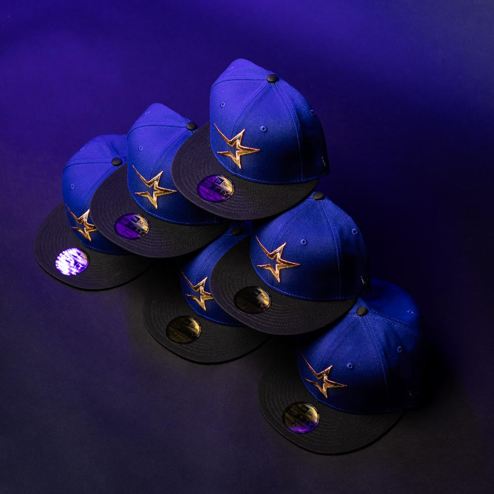 Six blue and black H Town North Houston Astros New Era Hat 5950 Fitted caps, each featuring a gold star logo inspired by the OG Dark Royal design, are arranged in a staggered pattern on a purple background, with a reflective sticker on the brim of each hat.