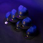 Six blue and black H Town North Houston Astros New Era Hat 5950 Fitted caps, each featuring a gold star logo inspired by the OG Dark Royal design, are arranged in a staggered pattern on a purple background, with a reflective sticker on the brim of each hat.