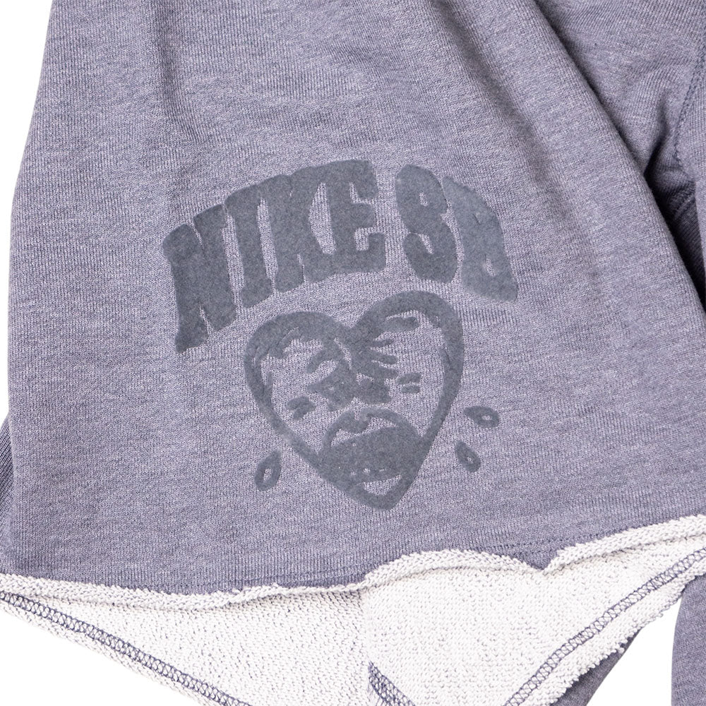 A close-up of IF1602065 Gray Sweat Short Skate Shorts by Nike SB shows light gray fabric with a faded SB logo above a sad, heart-shaped face and visible inner stitching at the loose bottom edge.