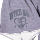A close-up of IF1602065 Gray Sweat Short Skate Shorts by Nike SB shows light gray fabric with a faded SB logo above a sad, heart-shaped face and visible inner stitching at the loose bottom edge.