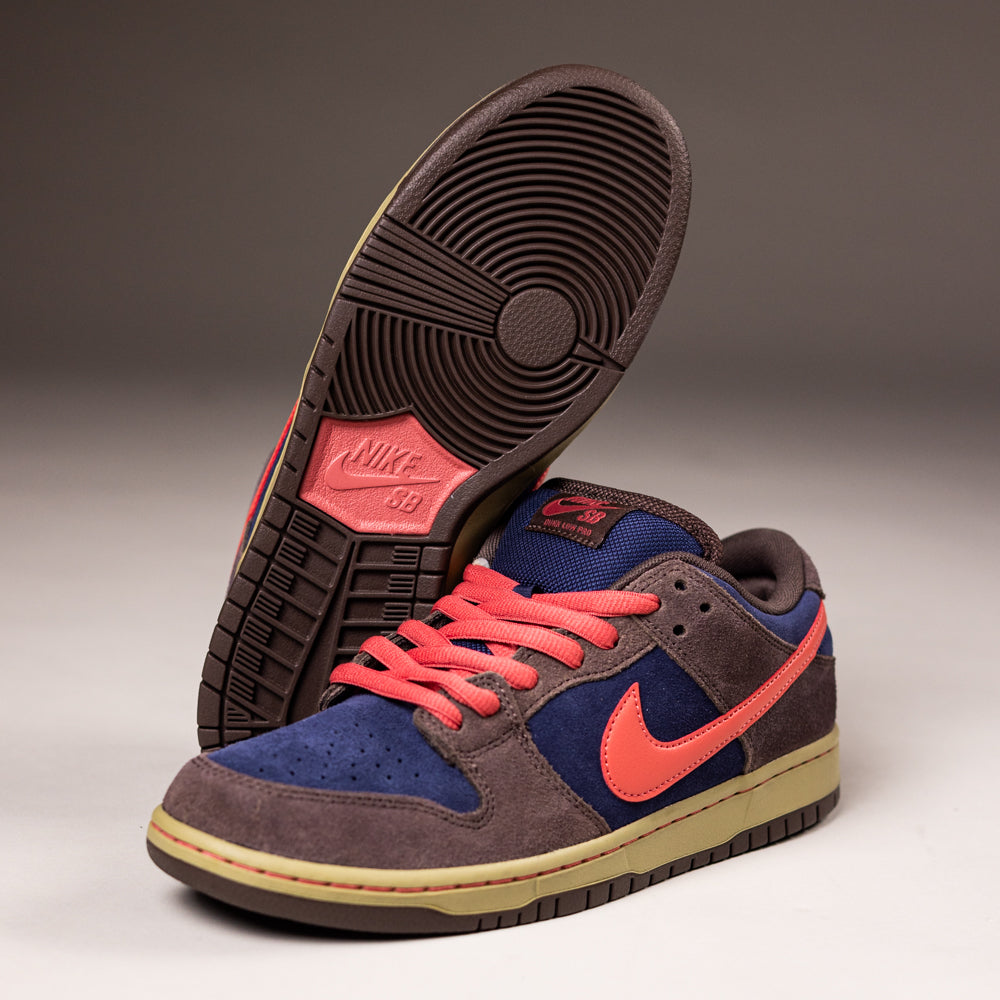 A pair of Nike SB Dunk Low Pro "Baroque Brown Adobe Midnight Navy" HQ1625200 suede shoes with dark brown, navy accents, bright coral laces and Swoosh, plus a beige sole. One shoe stands upright; the other displays its sole.