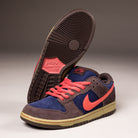 A pair of Nike SB Dunk Low Pro "Baroque Brown Adobe Midnight Navy" HQ1625200 suede shoes with dark brown, navy accents, bright coral laces and Swoosh, plus a beige sole. One shoe stands upright; the other displays its sole.