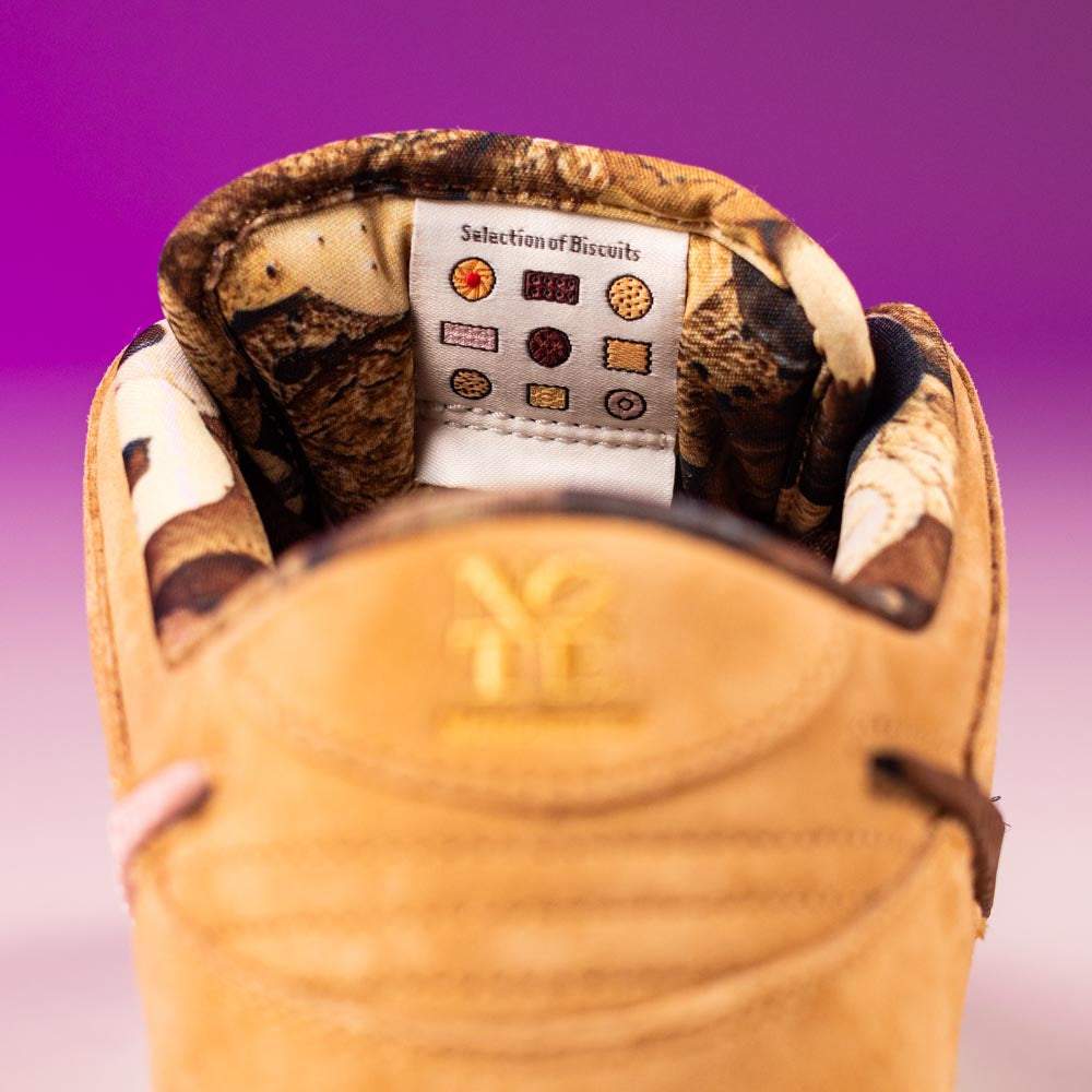 Close-up of the tan READ BELOW the BUTTON RAFFLE Nike SB Brew and Biscuits Dunk Low sneaker tongue, featuring a patterned lining and a white label with biscuit illustrations under "Selection of Biscuits" text. Background: purple-pink gradient.