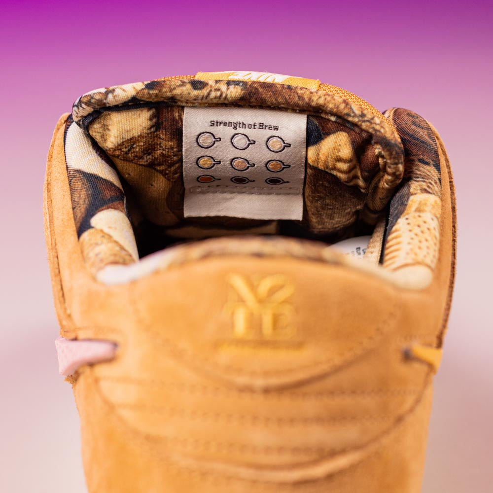 Close-up of the inner heel of the READ BELOW the BUTTON RAFFLE Nike SB Brew and Biscuits Dunk Low, featuring a patterned lining and a tag with colored dots reading "strength of Brave," set against a purple gradient background.