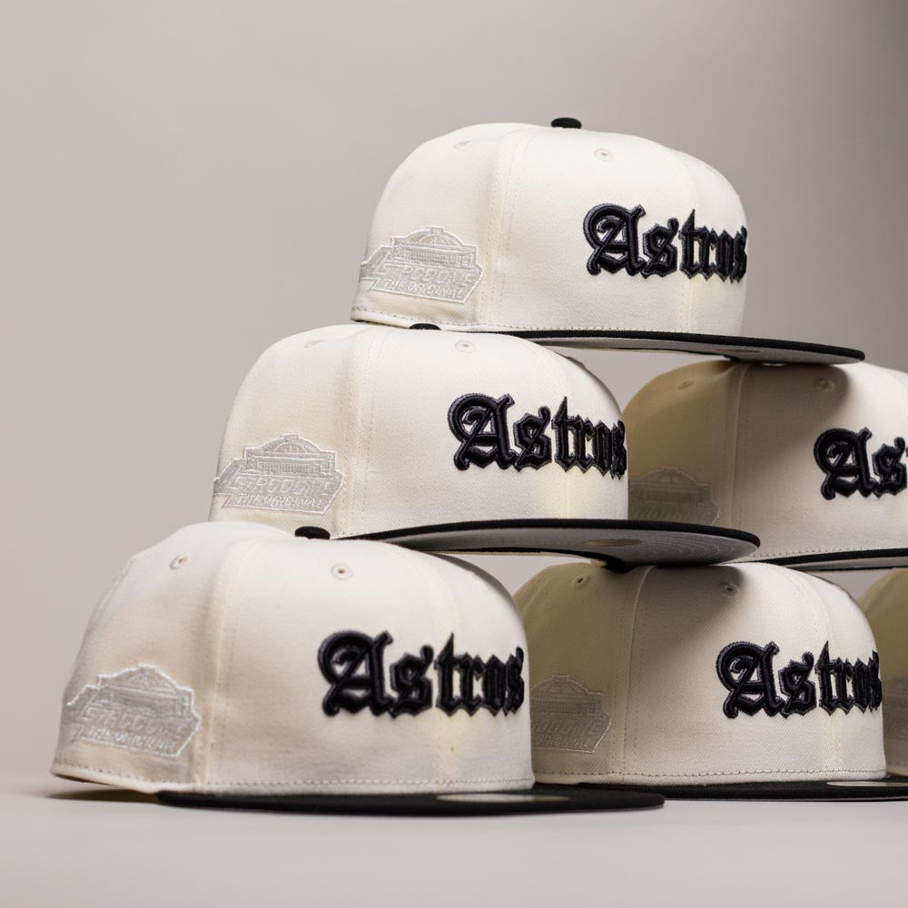 Six Straight Outta Southside OG Chrome Black Houston Astros New Era Hat 5950 Fitted caps, featuring cream-colored panels, black brims, and bold black Gothic “Astros” lettering, are stacked in a pyramid shape against a plain background. Each cap showcases the classic New Era 5950 Fitted style for a timeless look.