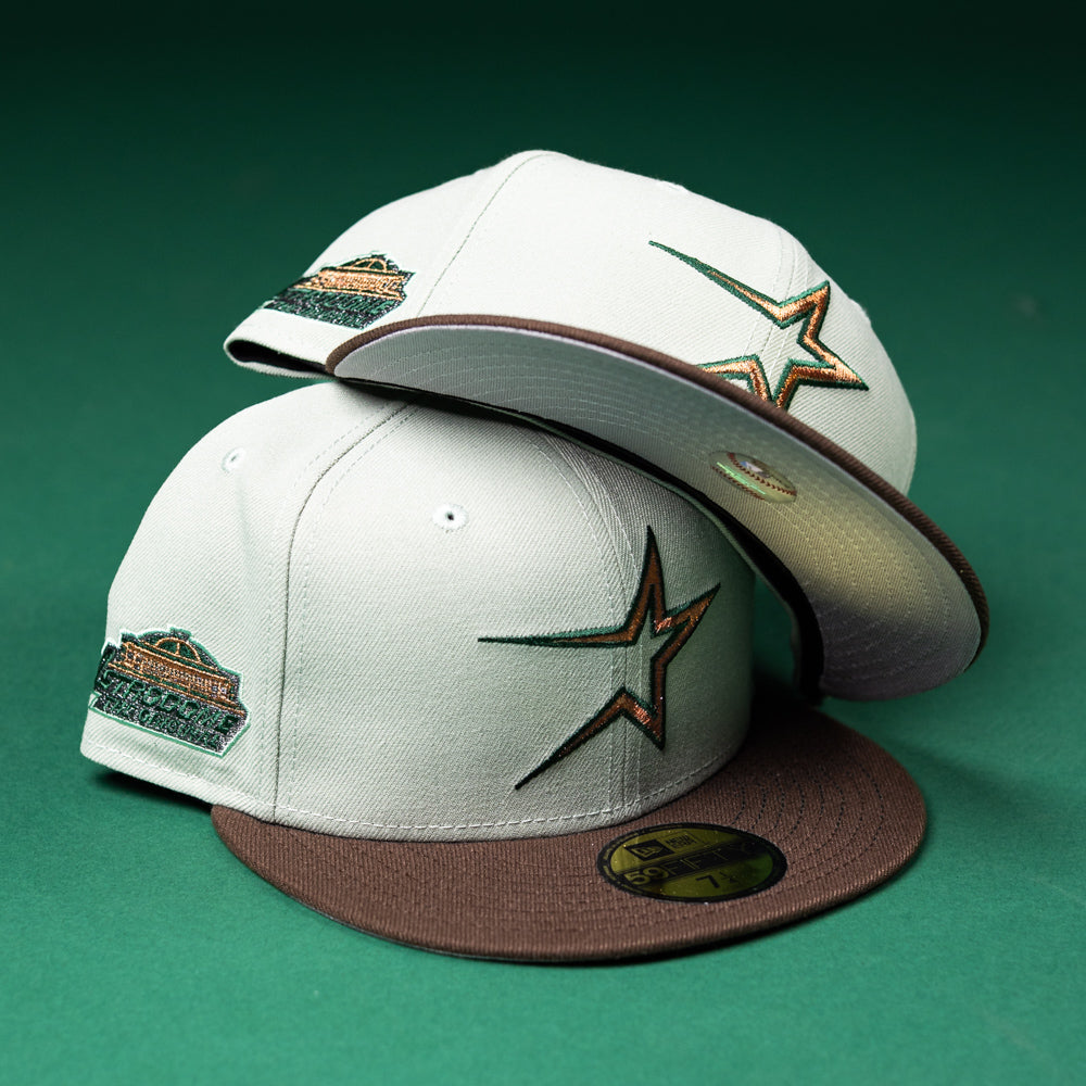 Two Copper Trails OG Walnut Egreen Houston Astros New Era 5950 fitted hats are displayed on a green background—one hat lying flat and the other leaning behind it—to showcase their white and brown color scheme, large embroidered star logos, commemorative patches, and distinctive Chrome Black accent.