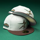 Two Copper Trails OG Walnut Egreen Houston Astros New Era 5950 fitted hats are displayed on a green background—one hat lying flat and the other leaning behind it—to showcase their white and brown color scheme, large embroidered star logos, commemorative patches, and distinctive Chrome Black accent.