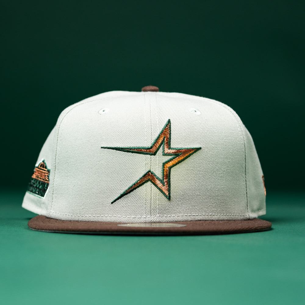 A Copper Trails OG Walnut Egreen Houston Astros New Era Hat 5950 Fitted, featuring a white crown, brown brim, and a stylized multicolor star logo on the front reminiscent of the classic Astros design, is displayed on a green surface with a green background.