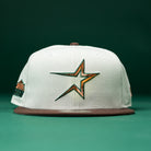 A Copper Trails OG Walnut Egreen Houston Astros New Era Hat 5950 Fitted, featuring a white crown, brown brim, and a stylized multicolor star logo on the front reminiscent of the classic Astros design, is displayed on a green surface with a green background.