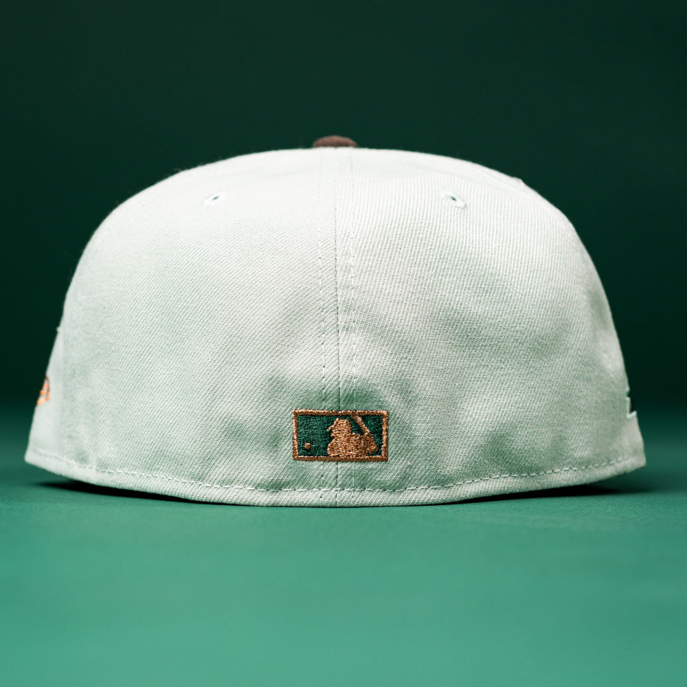 A Copper Trails OG Walnut Egreen Houston Astros New Era Hat 5950 Fitted is shown from the back, featuring a small MLB logo patch at the bottom center. Both the background and surface are green.