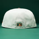 A Copper Trails OG Walnut Egreen Houston Astros New Era Hat 5950 Fitted is shown from the back, featuring a small MLB logo patch at the bottom center. Both the background and surface are green.
