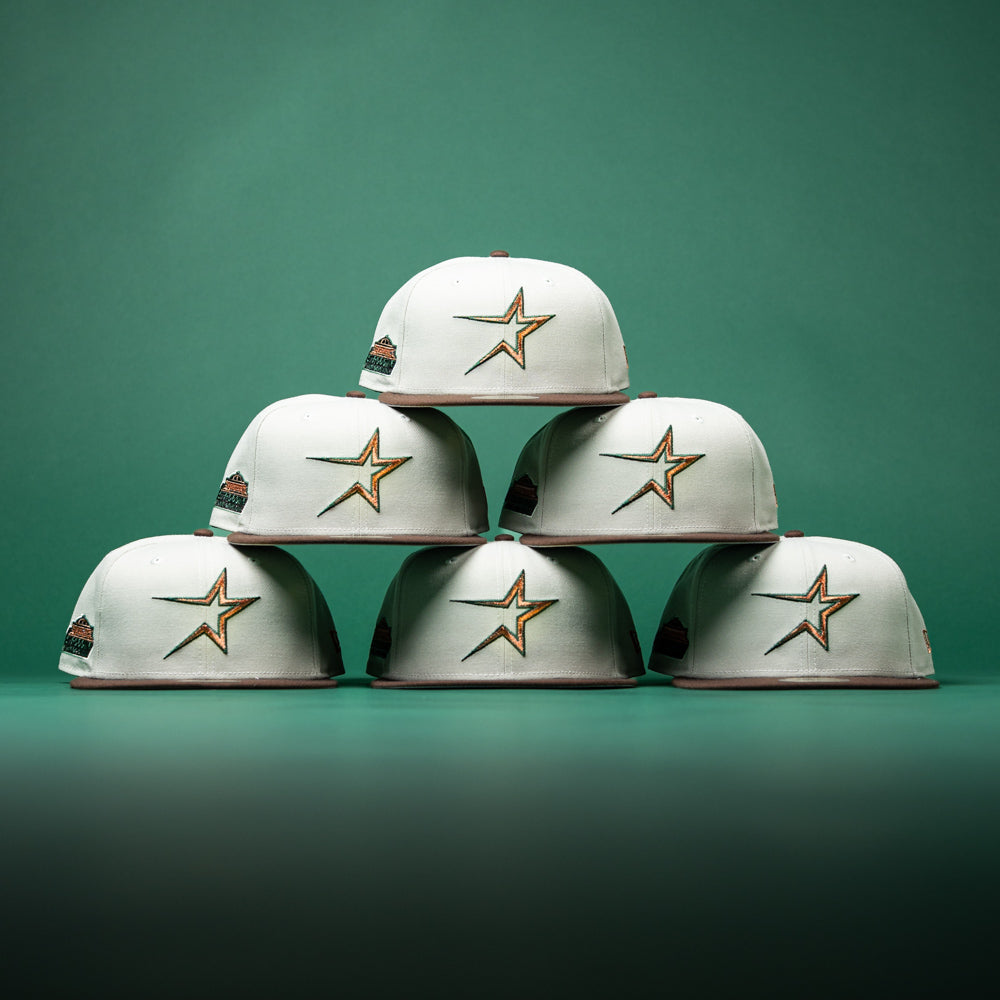 Six Copper Trails OG Walnut Egreen Houston Astros New Era 5950 Fitted hats are stacked in a pyramid shape against a green background.
