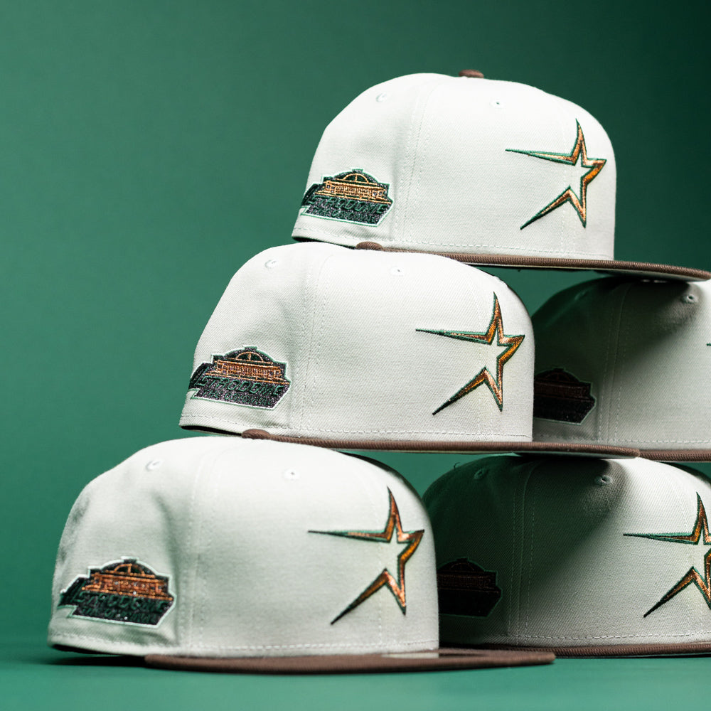 Six Copper Trails OG Walnut Egreen Houston Astros New Era 5950 fitted hats are stacked in two rows against a green background. Each white cap features a brown brim, a star logo on the front representing the Houston Astros, and a patch on the side.