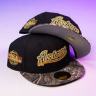 Two black Oil Spill WS05 Houston Astros New Era Hat 5950 Fitted caps by New Era, featuring gold embroidered logos, World Series patches, camouflage brims, and gold stickers, are displayed on a purple gradient background.