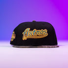 The Oil Spill WS05 Houston Astros New Era Hat 5950 Fitted features gold and yellow Astros embroidery on black, showcased on a flat surface with a purple gradient background—ideal for any Houston Astros fan.