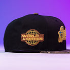 The Oil Spill WS05 Houston Astros New Era Hat 5950 Fitted is a black fitted cap featuring a golden World Series globe patch on the side and a partially embroidered Astros logo on the front, shown against a purple gradient background.