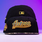 Two Oil Spill WS05 Houston Astros New Era 5950 Fitted hats with gold embroidery are stacked. The top cap features "Astros" in cursive; the back shows a gold MLB logo. The background shifts from purple to blue.