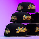 Four Oil Spill WS05 Houston Astros New Era Hat 5950 Fitted caps are stacked pyramid-style against a purple background, each featuring a gold World Series patch and a yellow Astros logo on the front.