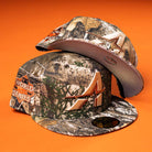 Two Panhandle Real Tree Orange Houston Astros New Era 5950 Fitted hats are displayed on an orange background—one upright, the other leaning to showcase their details.