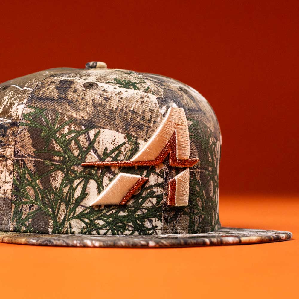 The Panhandle Real Tree Orange Houston Astros New Era 5950 Fitted hat features a camouflage pattern and an embroidered orange and tan stylized A logo on the front, shown against an orange background.