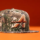 The Panhandle Real Tree Orange Houston Astros New Era 5950 Fitted hat features a camouflage pattern and an embroidered orange and tan stylized A logo on the front, shown against an orange background.