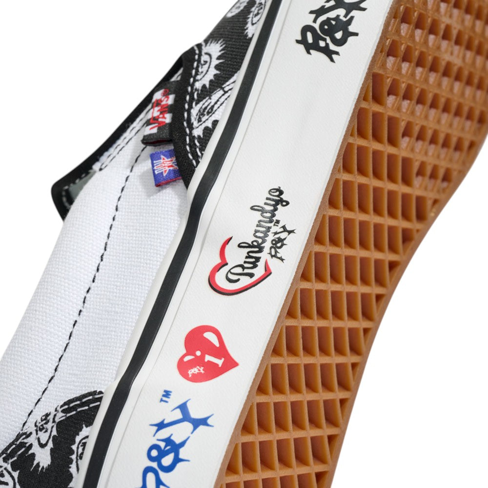 Here’s your rewritten sentence using the provided product data: Close-up of the Punk and Yo x Vans Skate Slip On VN000EKPY281 Canvas Shoe in white, featuring a brown rubber sole. This collaborative sneaker showcases black and red graphic designs and multilingual stylized text on the side—with details like a red heart—paying bold tribute to skateboarding streetwear culture.