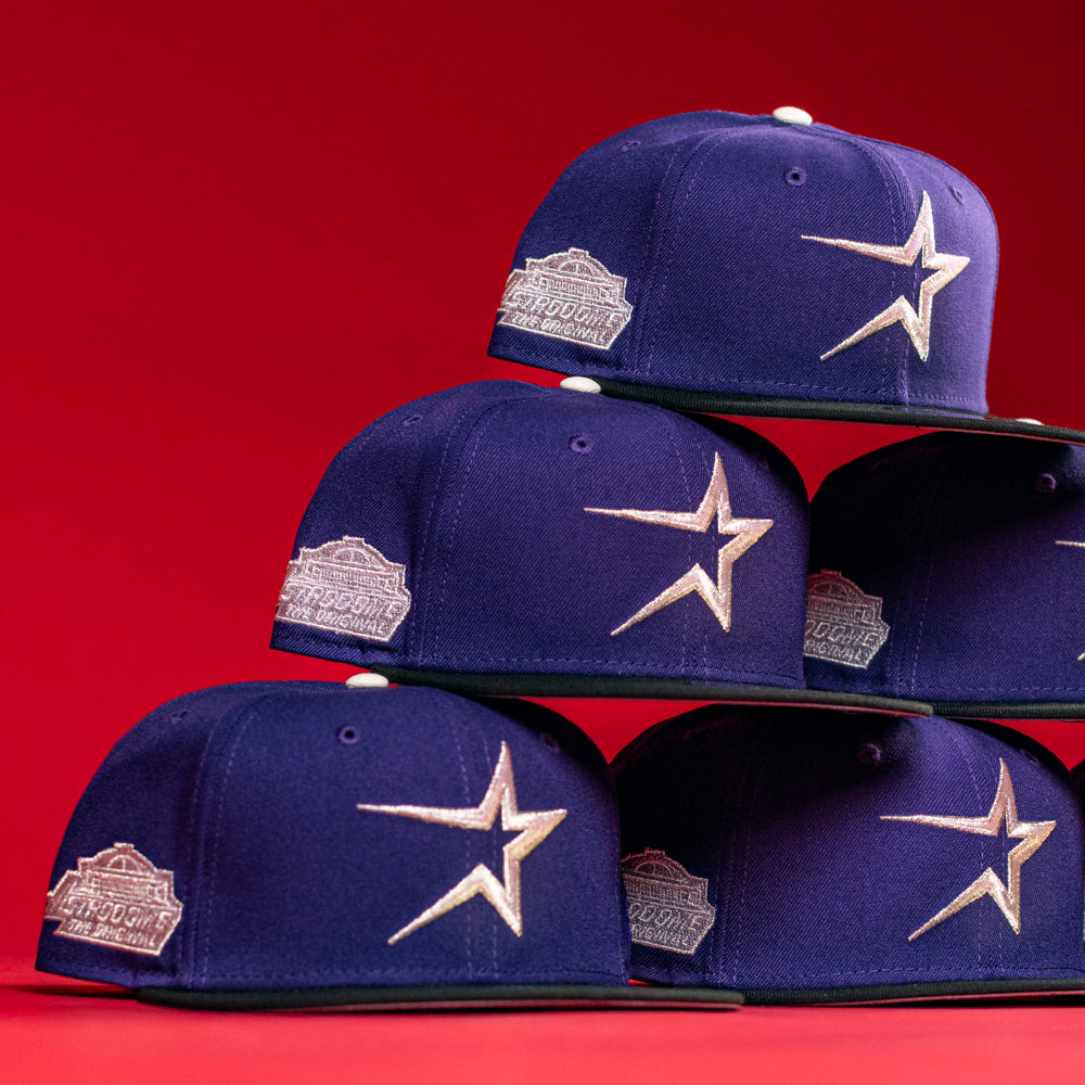 Six De Uva New Era Purple Houston Astros 5950 Fitted hats are stacked in a pyramid against a vibrant red background—a striking tribute to classic Astros style by BuryMeInTheSouf.