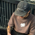 A tattooed person sits outdoors, wearing the Razor Arch Strip Hat Shadow Snapback from Independent Skateboards and a loose brown T-shirt with white text. They look down, partially obscuring their face.
