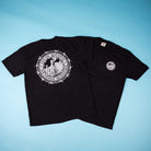 Two black Refinery Southside T-Shirts by Bigo are shown on a blue background: one has a large industrial graphic with text and building, the other features a smaller version on the left chest, both inspired by Southside Skatepark.
