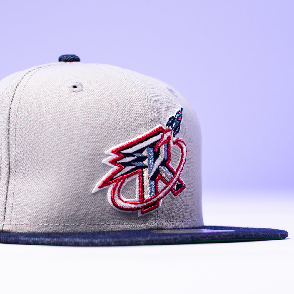A close-up of The Unit 95 ASG Denim Houston Rockets New Era Hat 5950 Fitted, a grey cap with black brim and a colorful red, white, and blue embroidered bird and letter K logo on the front, set against a purple background.
