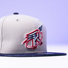 A close-up of The Unit 95 ASG Denim Houston Rockets New Era Hat 5950 Fitted, a grey cap with black brim and a colorful red, white, and blue embroidered bird and letter K logo on the front, set against a purple background.