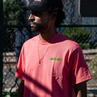 A man wearing a black cap and a Roskopp Cruz Control Heavyweight T-Shirt Coral Santa Cruz Tee stands outdoors in the sunlight next to a chain-link fence.