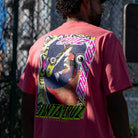 A man outdoors wears a Roskopp Cruz Control Heavyweight T-Shirt Coral Santa Cruz Tee featuring bold, colorful cartoon faces and the iconic Santa Cruz Skateboards logo on the back, with a wire fence in the background.